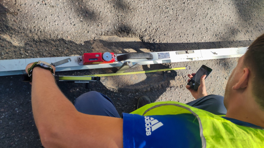 На асфальте лежит измерительная рейка с прибором, а дорожник фиксирует результаты на смартфон