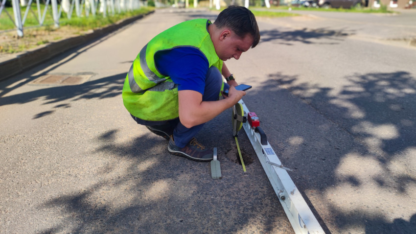 Рабочий присел у рейки и клин-промерника, снимая показания по неровностям асфальта