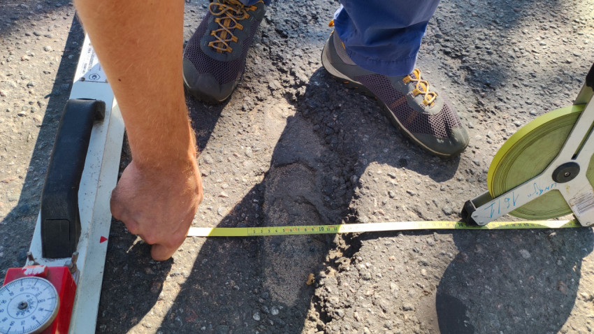 Рулеткой снимают ширину повреждения, а рядом лежит рейка для замера продольной ровности покрытия