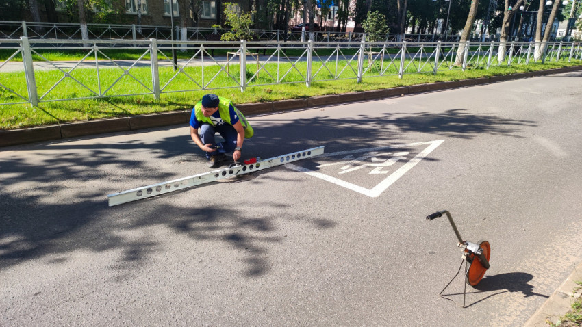 Дорожник присел на асфальт и проверяет разметку длинной рейкой рядом с измерительным колесом