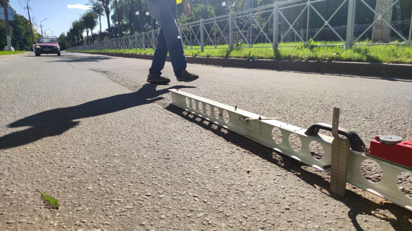 Длинная металлическая рейка и измерительный клин лежат на асфальте посреди улицы