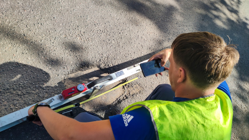 Дорожник снимает на телефон замер неровности: рейка, рулетка и прибор на асфальте