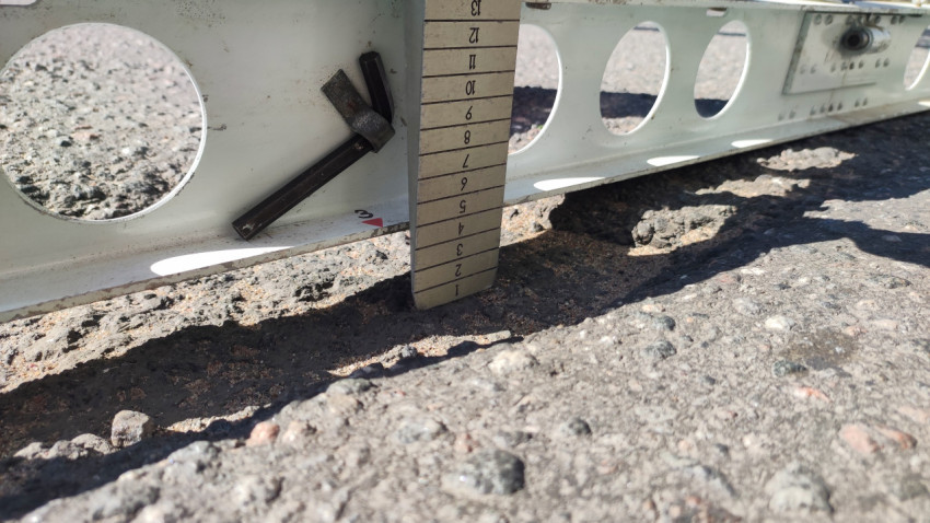 Крупный план замера глубины выбоины: клин-мерка под рейкой и крошка асфальта вокруг