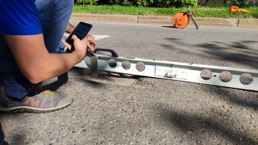 Крупный план замера глубины колеи: рейка на асфальте, клин-мерка и телефон в руках