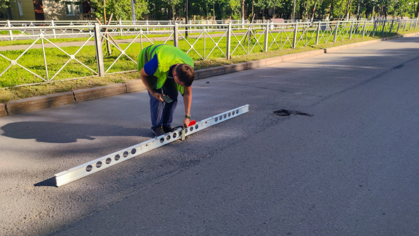 Парень в жилете наклонился над выбоиной и выставляет рейку для замера на дороге