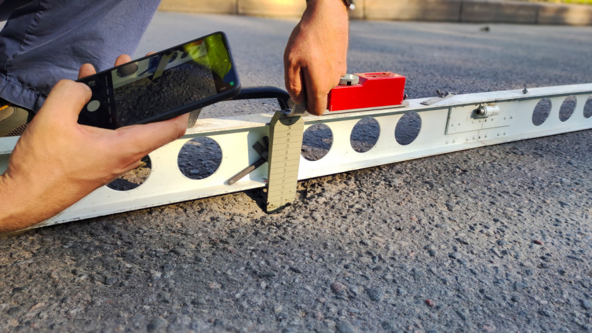 Руки держат телефон и клин у рейки, идёт фотофиксация замера неровности асфальта