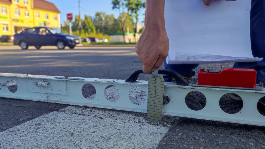 Рука с чертежами удерживает клин-измеритель под рейкой на полосе пешеходной разметки