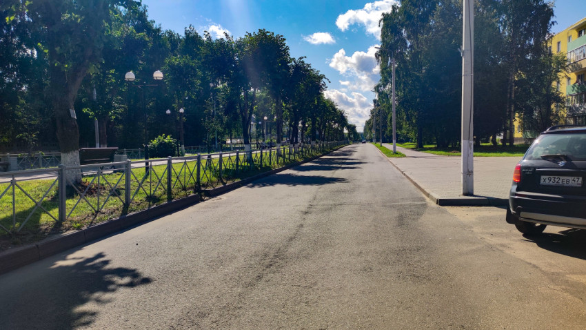 Прямая городская улица вдоль парка с ограждением, тротуаром и припаркованным внедорожником