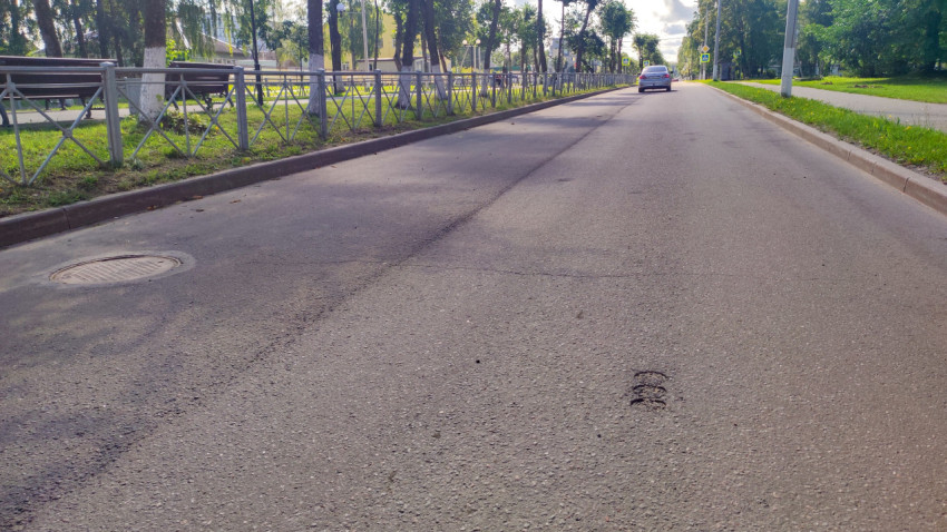 Проезжая часть с люком колодца, швом в асфальте и ограждением вдоль тротуара и зелёной зоны