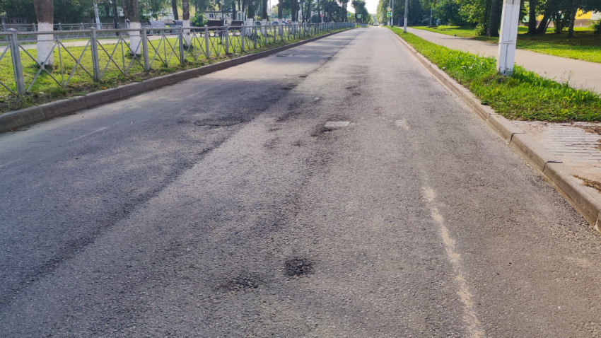 Пустая городская улица с асфальтовыми заплатками, бордюром и ограждением вдоль зелёной зоны