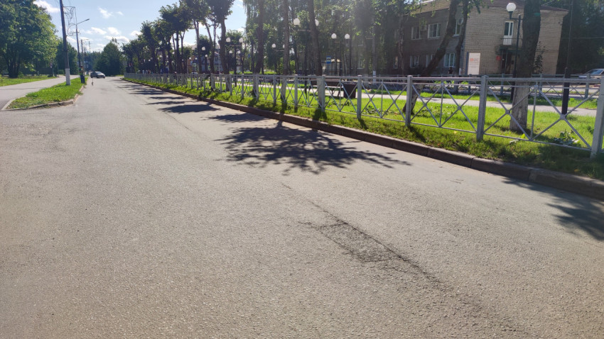 Заплатка на асфальте у ограждения и деревьев, длинные тени на почти пустой дороге