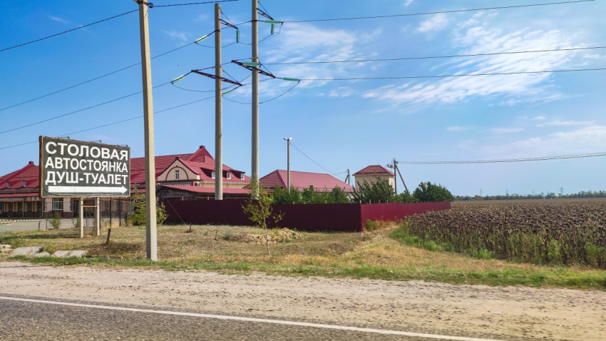 Придорожная вывеска «Столовая, автостоянка, душ-туалет» рядом с домами и полем