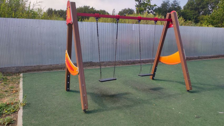 Пустые качели на цепях на зелёном покрытии у металлического забора