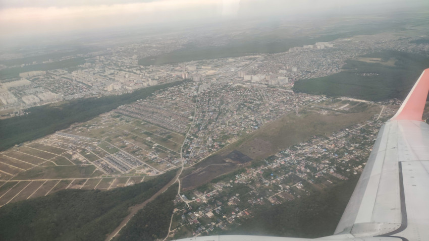 Вид из иллюминатора: крыло самолёта над городскими кварталами и лесом