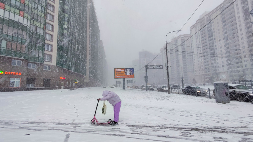 Человек с самокатом на фоне плотного снегопада и высотной застройки