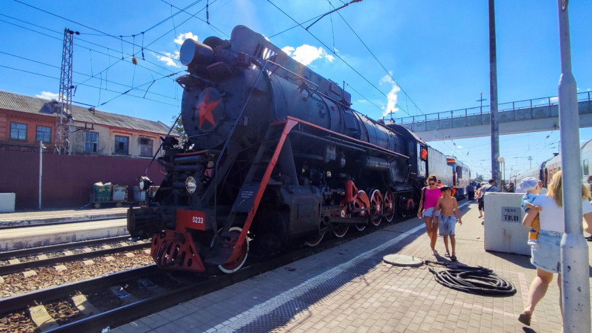 Паровоз на путях станции с посетителями, контактной сетью и кабельными бухтами на перроне