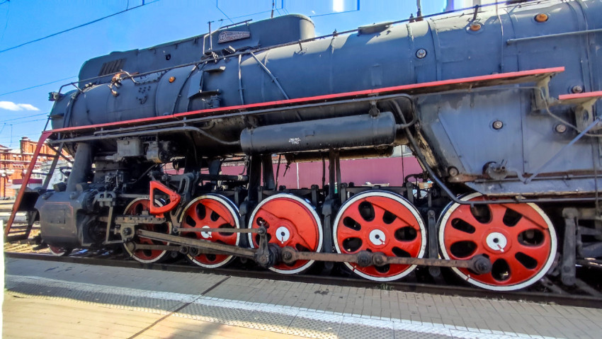 Боковой фрагмент паровоза с котлом и красными ведущими колёсами на железнодорожной станции