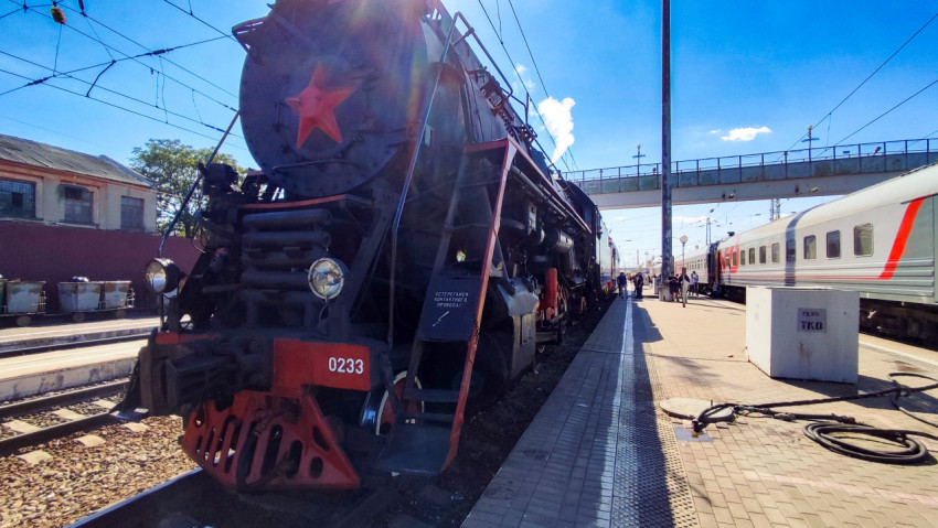 Паровоз ЛВ-0233 у платформы с перспективой путей, пешеходным мостом и солнечными бликами