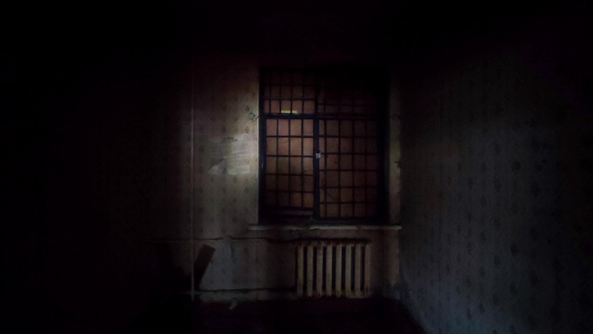 Окно с решёткой и радиатор, снятые через тёмный проём в заброшенной комнате