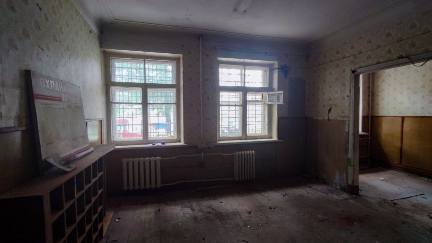 Пустая комната с окнами в решётках, радиаторами и стендом «Охрана труда»