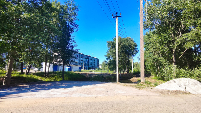 Перекрёсток с участком отсыпанного основания и опорами ЛЭП на фоне жилого дома и деревьев