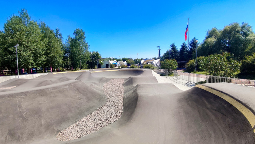 Волнообразная трасса памп-трека с гравийной дренажной полосой на фоне деревьев и флага