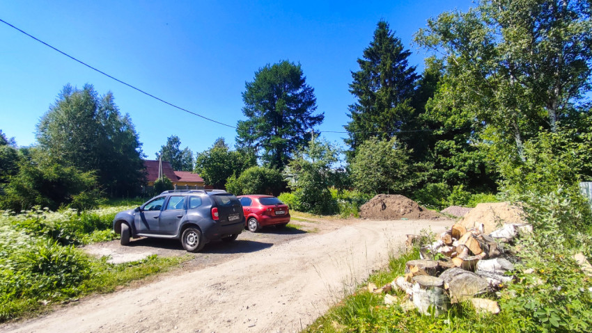 Грунтовая дорога с припаркованными автомобилями, кучами песка и поленницей у обочины