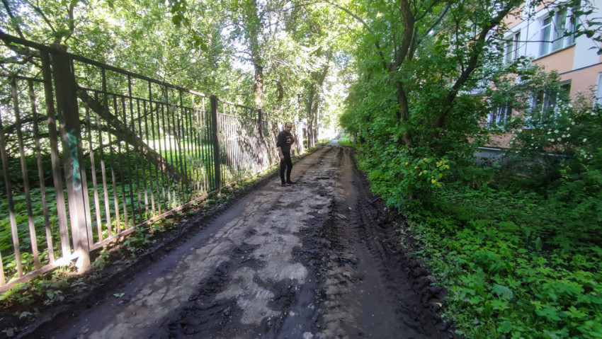 Грязная дворовая дорога со следами техники вдоль забора и зелёных насаждений