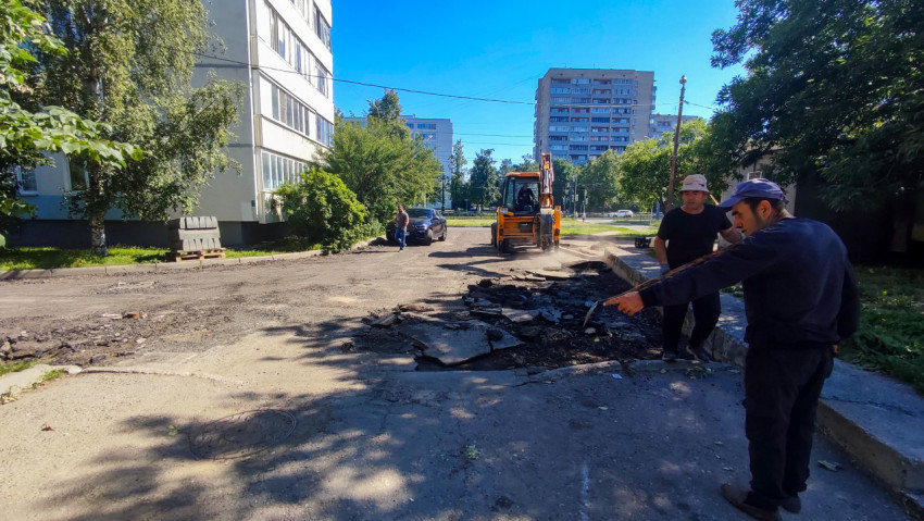 Экскаватор-погрузчик и рабочие разбирают асфальт во дворе жилого квартала