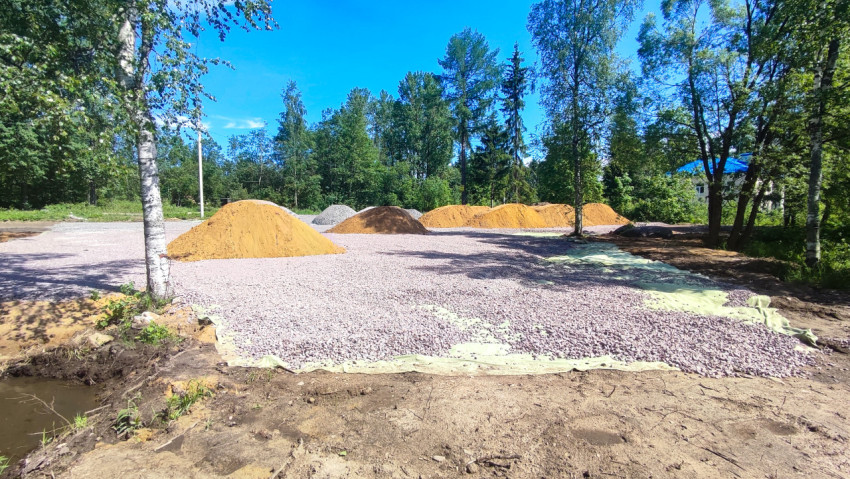 Отсыпанная щебнем площадка с кучами песка и берёзами, на заднем плане дом с синей крышей