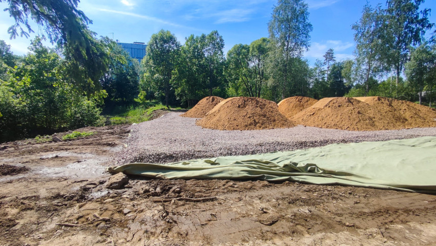 Слои щебня и геотекстиля с рядами песчаных куч на площадке у леса и здания