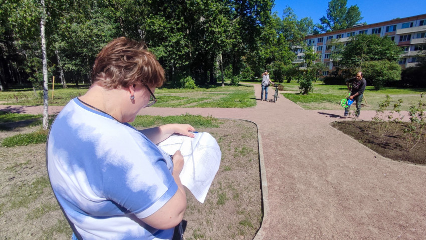 Женщина сверяет документы, пока рабочий с измерительным колесом идёт по дорожке в дворовом сквере