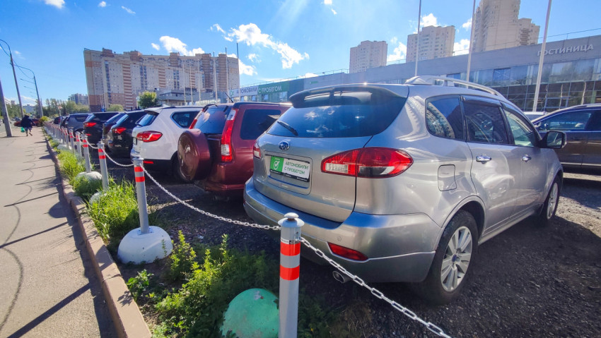 Ряд подержанных автомобилей у тротуара с цепным ограждением на фоне автосалона