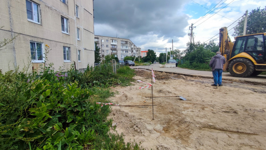Дворовая стройплощадка у жилого дома с песчаным основанием, колышками разметки и экскаватором-погрузчиком