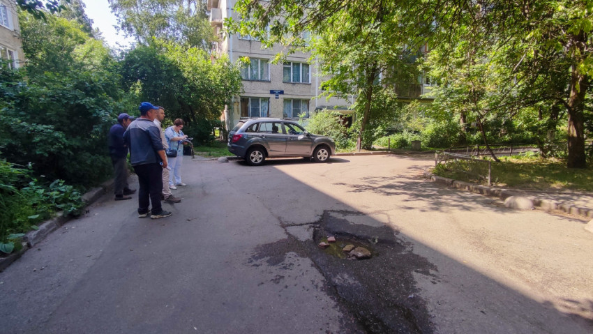 Яма с водой на дворовой дороге на фоне жилого дома и группы людей с документами