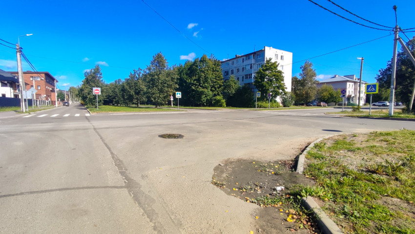 Перекрёсток с разметкой, дорожными знаками и участком грунта у бордюра на фоне жилых домов и деревьев