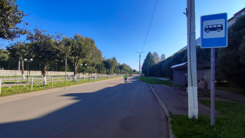 Знак автобусной остановки у павильона и пустая дорога, вдаль уходит человек с измерительным колесом