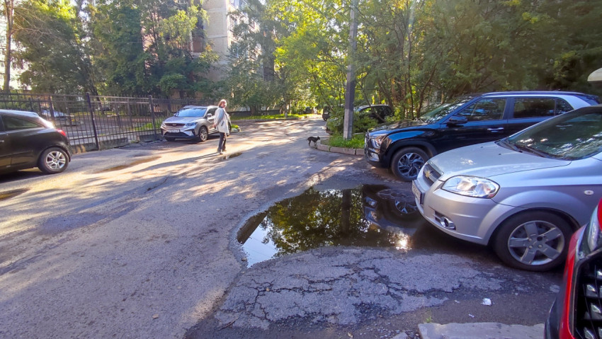 Дворовая парковка с рядом автомобилей, лужей и изношенным асфальтом у жилых домов