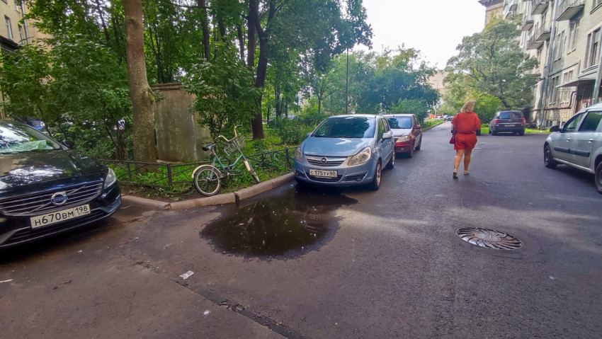 Дворовая дорога с лужей, велосипедом у ограждения и женщиной на фоне деревьев и припаркованных машин