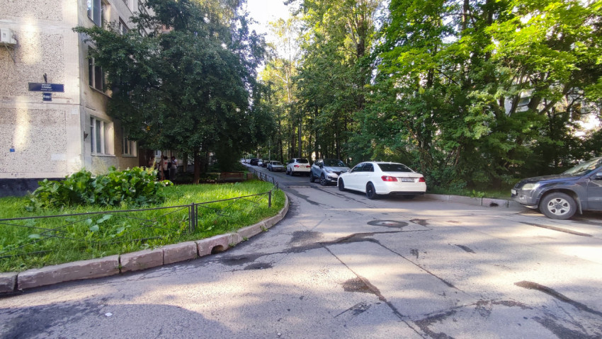 Поворот дворового проезда с припаркованными автомобилями, ограждением и зелёной зоной у многоэтажки