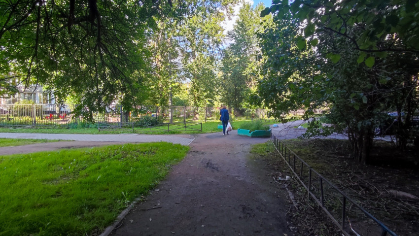 Утоптанная грунтовая пешеходная дорожка под кронами деревьев у металлического ограждения во дворе