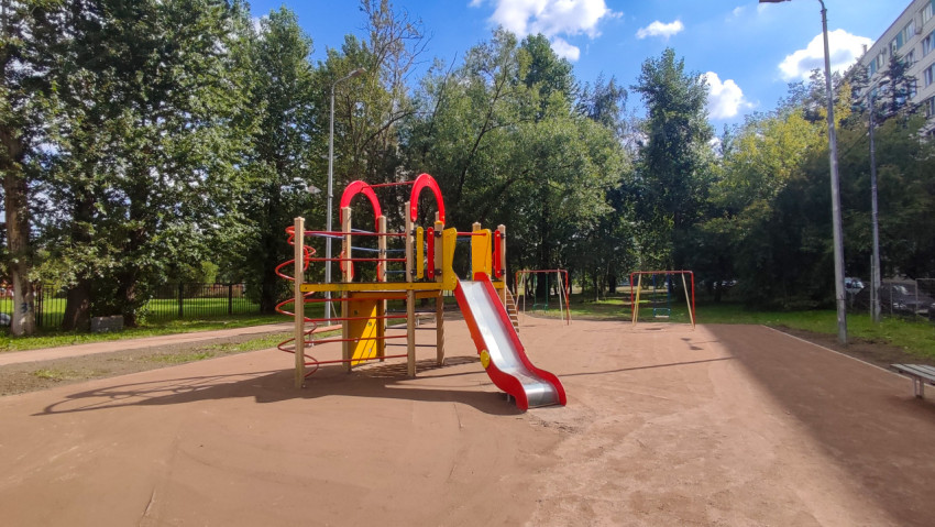 Детская площадка с горкой и качелями на песчаном основании среди деревьев