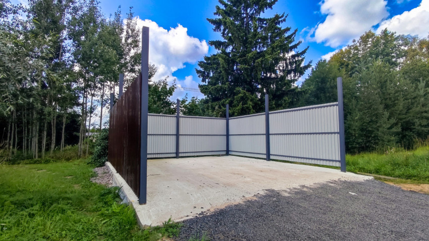 Металлическое ограждение из профнастила на бетонной площадке на фоне хвойных деревьев и облачного неба