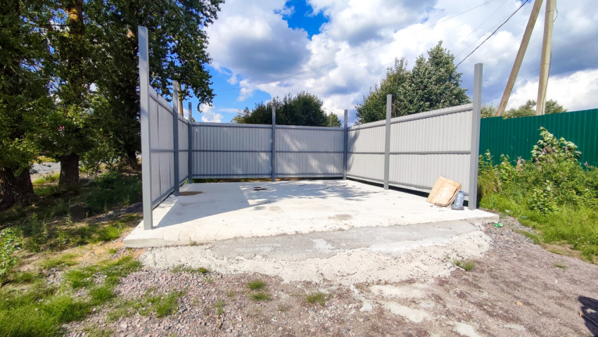 Открытая бетонная площадка с ограждением из серого профнастила, деревьями и линиями электропередачи