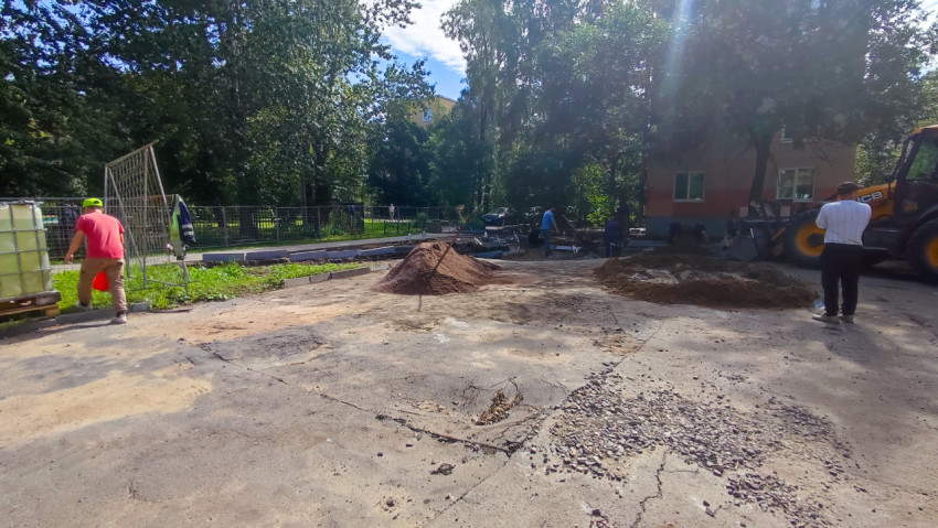 Благоустройство двора с экскаватором-погрузчиком, кучами песка и рабочими у разобранного покрытия