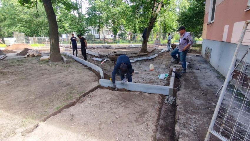Монтаж бордюров вдоль фасада здания с траншеей, щебнем и рабочими на придомовой территории