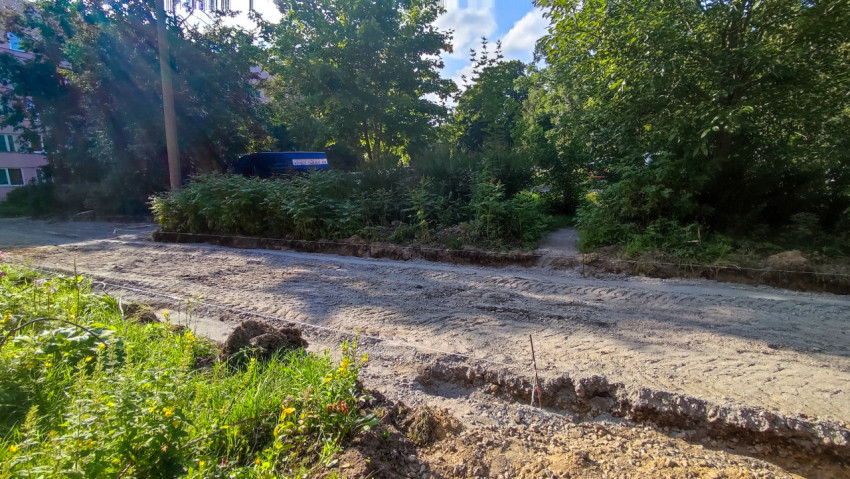 Выравненная песчано-грунтовая поверхность будущего проезда с колышками разметки рядом с зелёными кустами