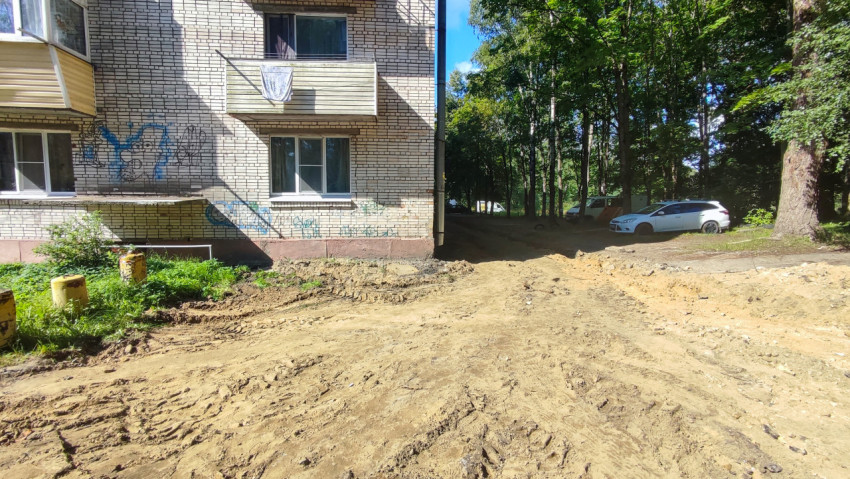 Выравненная песчаная площадка у угла кирпичного дома рядом с зелёной зоной и парковкой