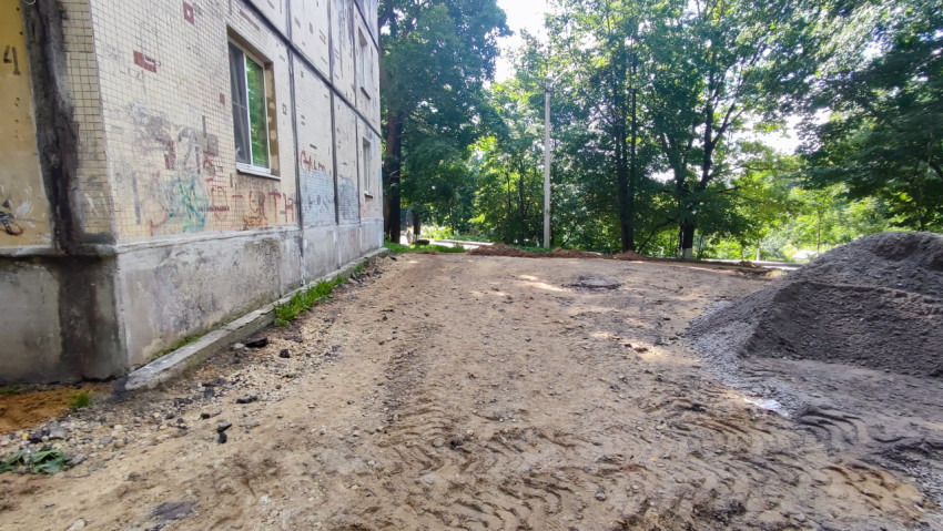 Песчаное основание дворового проезда у кирпичного дома с кучей щебня и следами техники