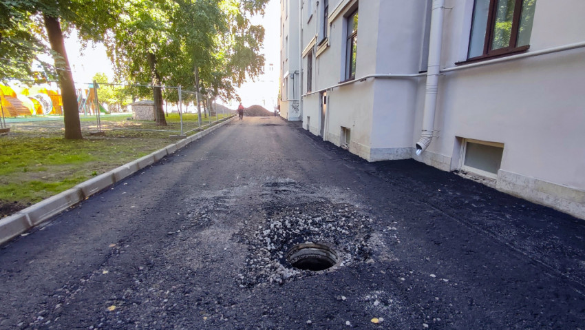 Открытый колодец без крышки на новом асфальте вдоль дома рядом с ограждением и деревьями